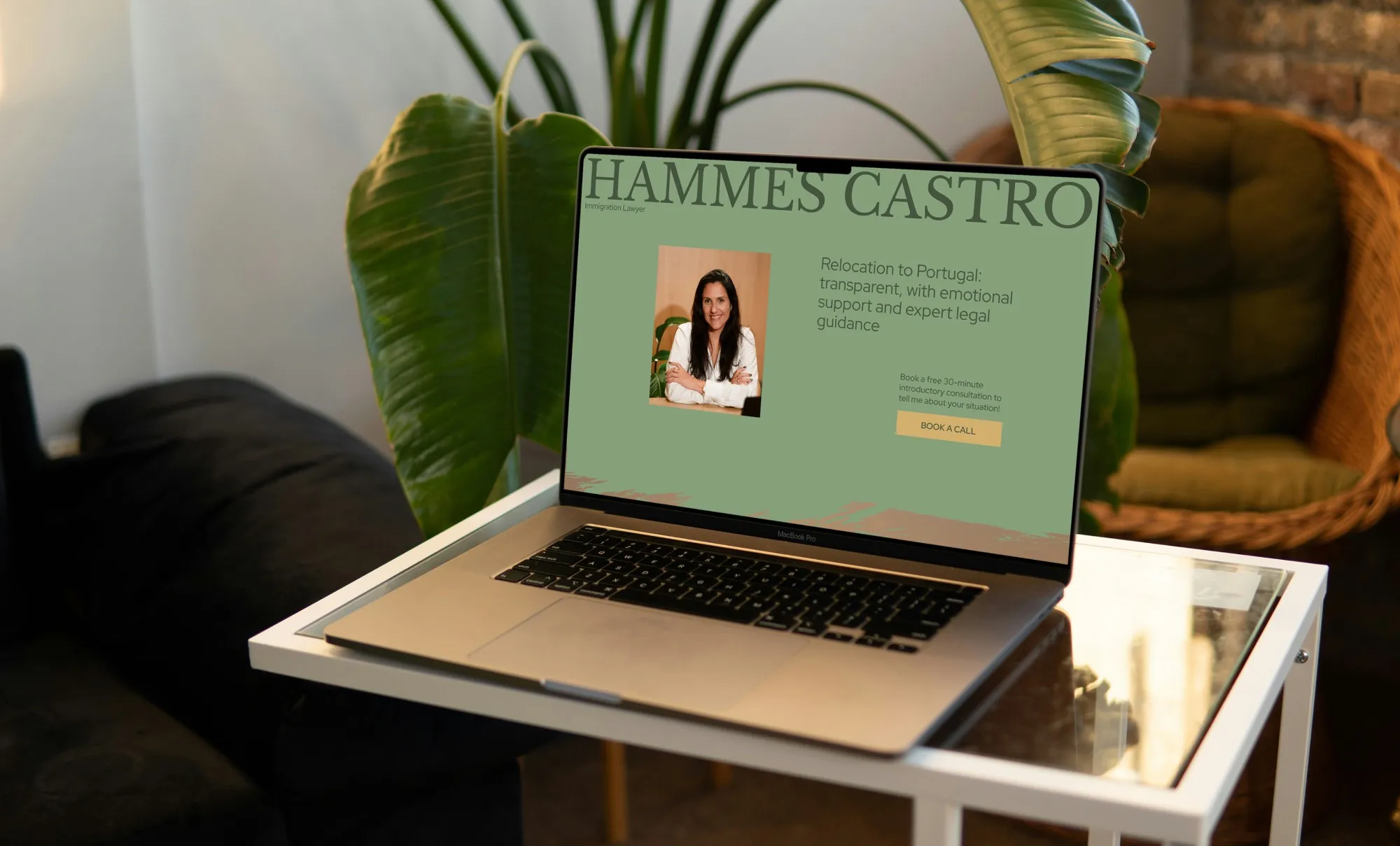Laptop on a glass side table beside a plant, showing the green Hammes Castro homepage: “Relocation to Portugal” and a Book a call button
